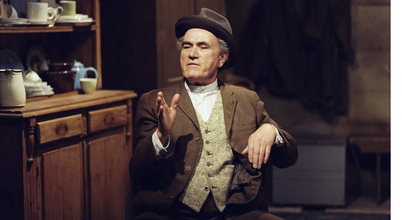Éamon Kelly seated in a rustic interior, gesturing as he tells a story.