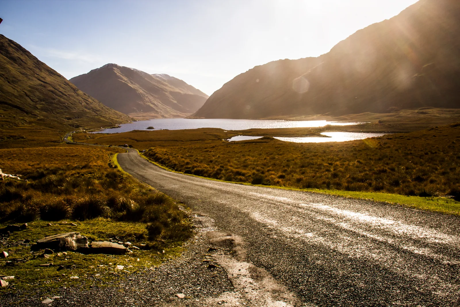 Golden sunlight falls across Doo Lough and the surrounding Mayo mountains, with a winding road in the foreground.