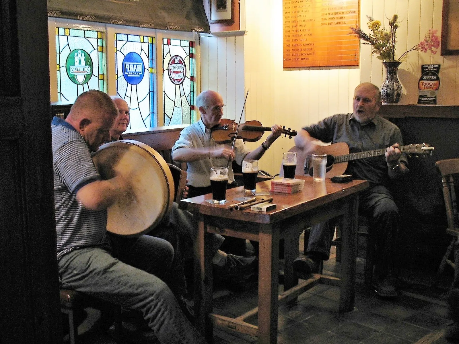 Musicians playing bodhrán, fiddle and guitar around a pub table.
